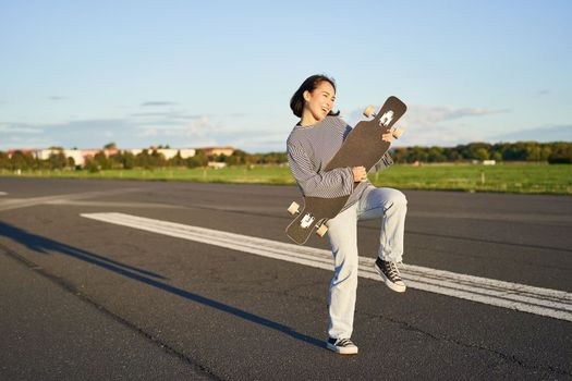 Happy brunette asian girl having fun on street with longboard, skating, using skateboard as guitar, enjoying skateboarding.