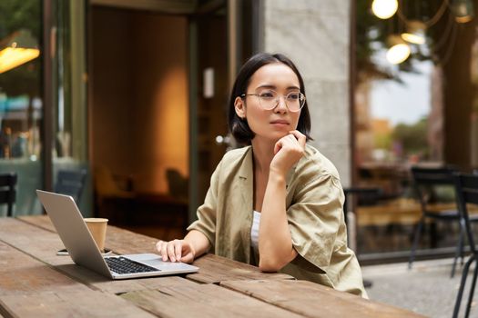 Working woman sitting with laptop in cafe and thinking. Asian girl in glasses works remotely, drinks coffee and looks thoughtful.