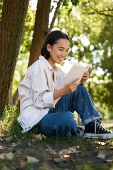 Vertical shot of happy asian woman relaxing outdoors in park, reading her book and sitting under tree shade on warm sunny day.