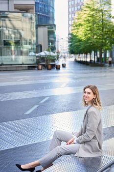 Vertical shot of smiling businesswoman in beige suit, sitting outside business office building, looking with confidence.