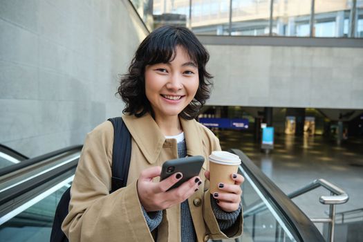 Portrait of beautiful brunette girl on escalator, drinks coffee to go and uses smartphone, smiles at camera, goes to work or univeristy, commutes.