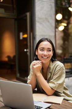 Portrait of asian girl works outdoors in cafe, sits with laptop, studies, smiles happy.