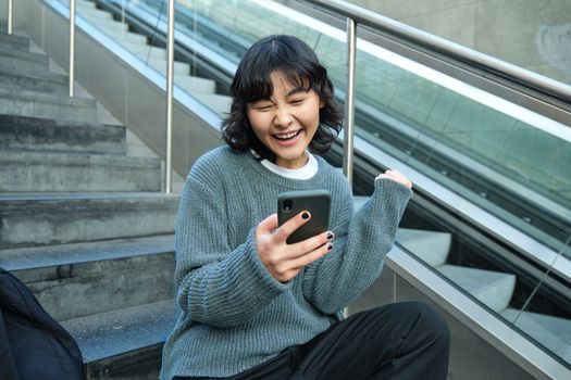 Portrait of brunette young woman, student sits on stairs in public place, looks at smartphone, reads text message and smiles.