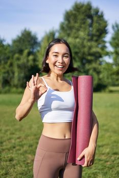 Excited young woman standing with sports mat, yoga clothes, shows okay sign, workout in park, wellbeing training session outdoors.