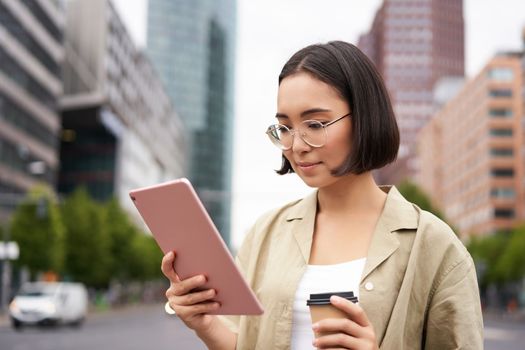 Portrait of happy young woman in glasses, standing on street with cup of coffee and tablet.