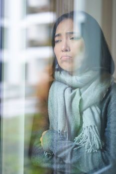 Portrait of sad ill korean woman looking out of the window, staying at home due to coronavirus, being sick and cant leave house.