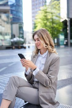 Smiling female employee in suit, sitting in city center and using mobile phone.