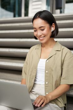 Remote worker. Smiling asian girl sits outdoors on street with laptop. Happy young woman working on computer remotely, listen music in wireless headphones.