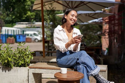 People and communication. Stylish asian girl sits in outdoor cafe with cup of coffee and smartphone, using mobile phone.