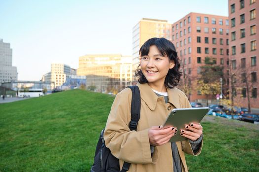 Happy young brunette girl, asian woman walks around city with tablet, goes to university with her digital gadget and backpack.