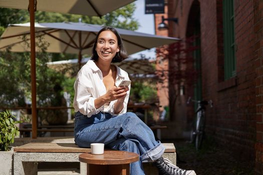 Stylish young asian woman sitting with cup of coffee in an outdoor cafe, smiling and looking happy, looking around.