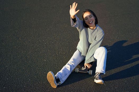 Positive korean girl covers her face from sunlight, sits on skateboard and smiles happily. Copy space