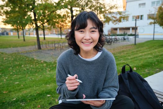 Young asian girl with graphic pencil and tablet, sits in park on bench, draws scatches, does her homework outdoors.