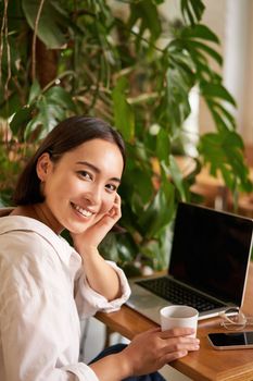 Trendy young woman sitting in cafe and smiling at camera, drinking coffee and using laptop, working remotely, studying or browsing internet.