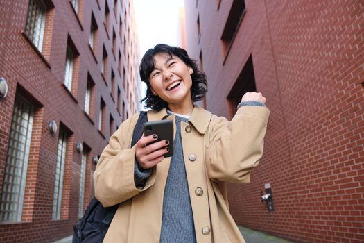 Enthusiastic young woman dances on street, celebrates victory, triumphs, holds smartphone, reads text message on mobile phone, walking in city.