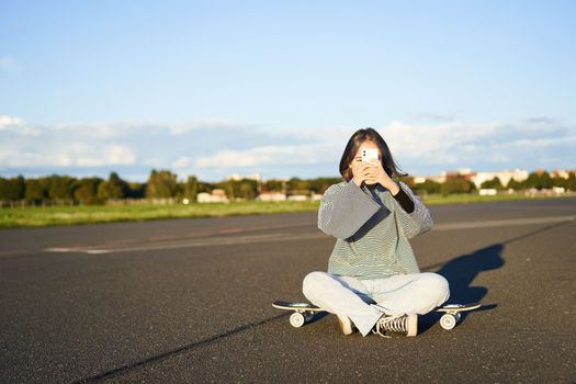 Hipster teen girl sitting on her skateboard, taking photos on smartphone. Asian woman skater sits on longboard and photographing on mobile phone.