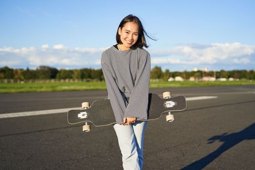 Portrait of beautiful asian girl skating on longboard, crusing with skateboard on empty road, enjoying freetime on fresh air.