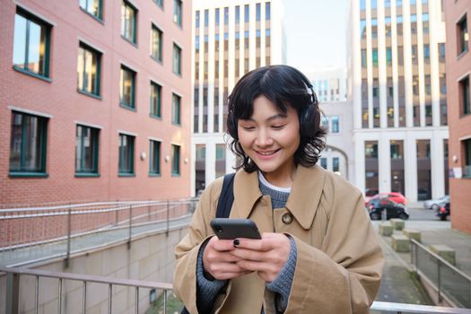 Portrait of cute girl tourist, korean woman in headphones, looks at mobile phone, uses smartphone app, map or text messages, listens music in headphones.