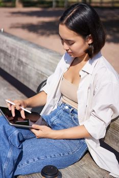 Young asian creative girl drawing with pen on graphic tablet, sitting outdoors in park, painting smth.