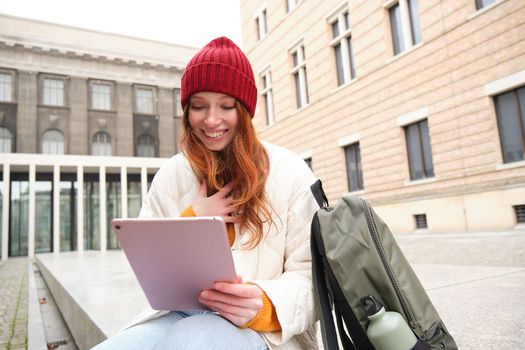 Redhead girl smiles, sits outdoors near building with digital tablet, thermos and backpack, connects to public internet and searches smth online on her gadget.