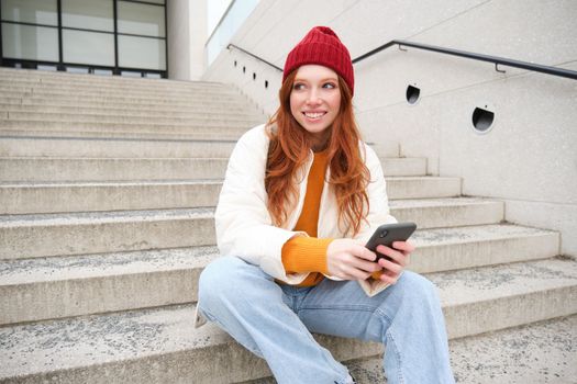 Redhead girl in red hat, sits on stairs and uses mobile phone. Modern woman holding smartphone, texting message, using telephone application outdoors.