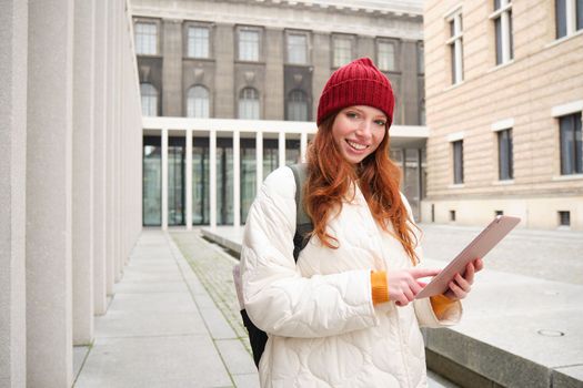 Stylish ginger girl, tourist walks with digital tablet around city, woman connects to iternet on her gadget, looking up information, texting message.