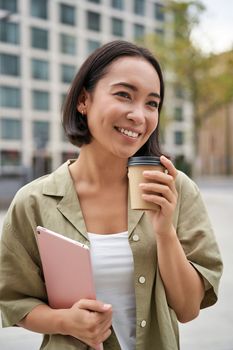 Vertical shot of beautiful korean girl drinks coffee, holds tablet, poses on street of city. Copy space