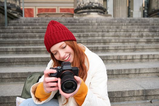 Portrait of young photographer girl, sits on stairs with professional camera, takes photos outdoors, making lifestyle shooting.