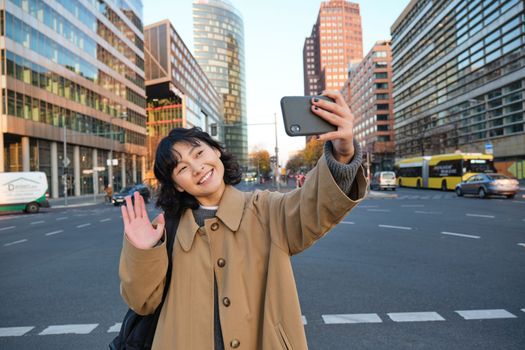 Happy asian girl tourist, takes selfie in city centre, makes video call and waves at smartphone camera, greets someone over phone.