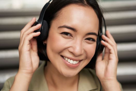 Close up portrait of smiling asian girl in headphones, listens to music outdoors, looking happy. People concept