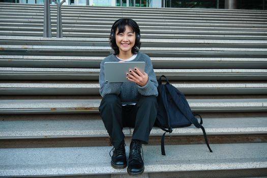 Young woman draws on her tablet, listens to music in headphones. Asian girl sits on stairs and does graphic design project, sits on street stairs.