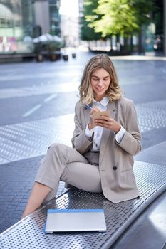 Businesswoman sitting with laptop, wearing suit, writing down notes in her planner, being in city centre outdoors.