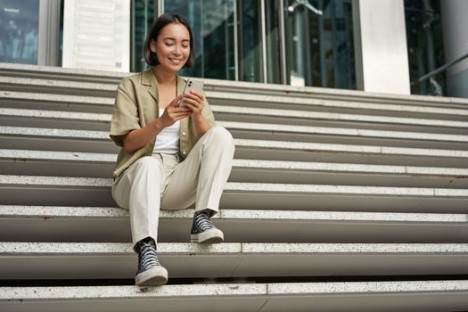 Cellular technology and people. Young happy asian girl sits with smartphone in front of building. Woman using mobile phone, smiling while looking at screen.