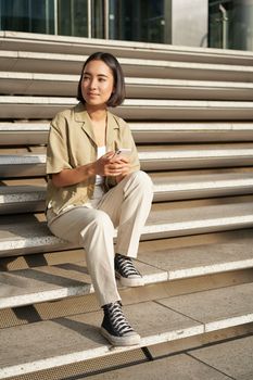 Beautiful asian girl sitting on stairs outside building, using mobile phone, looking at smartphone app and smiling.