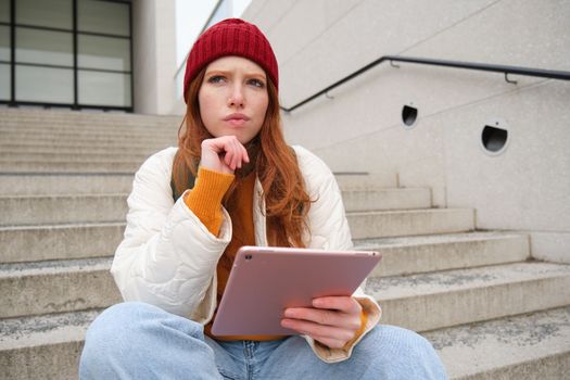 Portrait of troubled redhead girl, college student with thoughtful face, sits on street stairs, holds digital tablet, thinks how to reply on message.