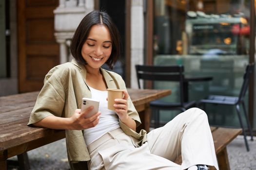 Stylish urban girl sits with her phone in cafe, drinks coffee and chatting, browsing social media on smartphone.