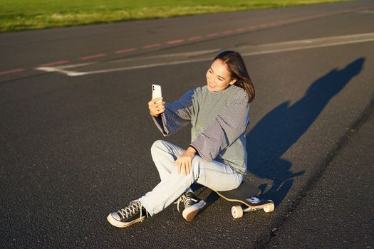 Beautiful korean girl takes selfie on smartphone, takes photo with her skateboard, enoys sunny day outdoors.