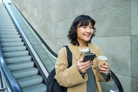 Portrait of smiling korean girl commutes, goes somewhere in city, drinks coffee to go and uses smartphone, stands on escalator.