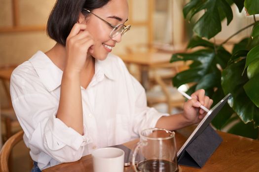 Portrait of beautiful young woman sitting in cafe, drinking coffee and drawing diagram on tablet with graphic pen, smiling pleased.