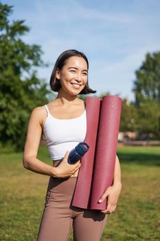 Portrait of young slim and healthy korean girl doing workout in park, standing with water bottle and rubber mat for execises on green lawn, smiling happily.