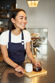 Vertical shot of smiling girl barista serving coffee, making batch brew, filter for client in cafe, wearing blue apron behind counter.