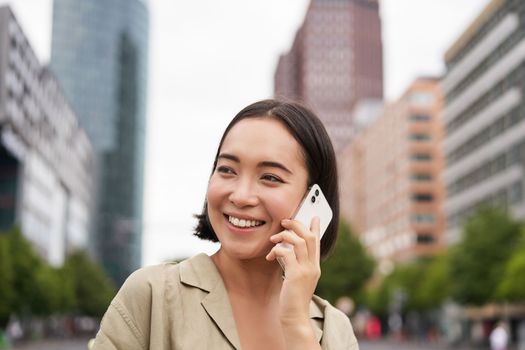 Portrait of smiling asian woman talking on mobile phone, walking on street near busy road and speaking to friend, laughing.