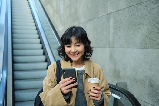 Portrait of smiling korean girl commutes, goes somewhere in city, drinks coffee to go and uses smartphone, stands on escalator.