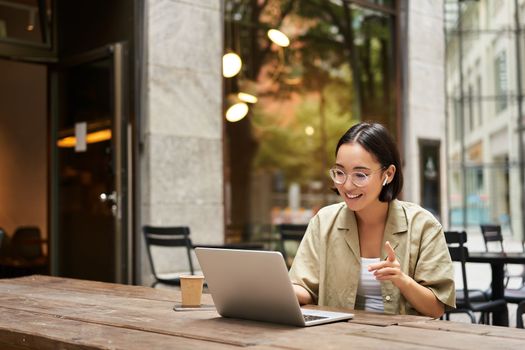 Young woman sitting on online meeting in outdoor cafe, talking to laptop camera, explaining something, drinking coffee.