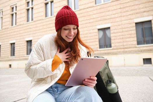 Beautiful redhead woman in red hat, sits with backpack and thermos, using digital tablet outdoors, connects to wifi, texts message, books tickets online.