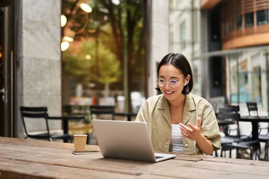 Young woman sitting on online meeting in outdoor cafe, talking to laptop camera, explaining something, drinking coffee.