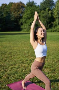 Vertical shot of young asian fitness girl doing yoga asana, standing in tree pose and smiling, execising in park on rubber mat.