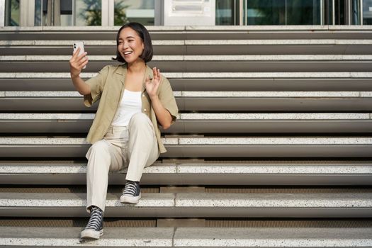 Portrait of asian beautiful girl takes selfie on smartphone. Young korean woman sits on stairs outdoors and makes photos on mobile phone.