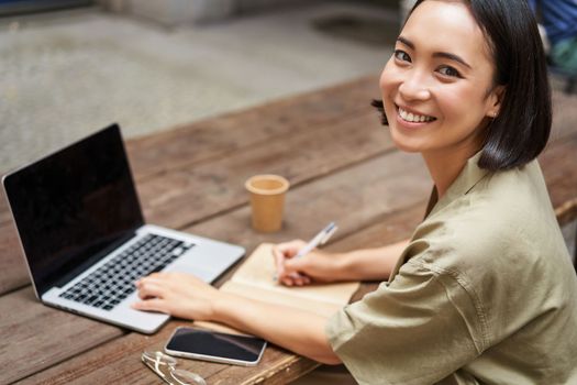 Portrait of young asian woman working on laptop, screen is blank. Girl working outdoors on remote, studying, e-learning and smiling.