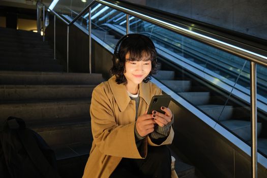 Technology. Portrait of asian girl sits on stairs near escalator, listens music in headphones and uses social media, reads, watches video in public place.